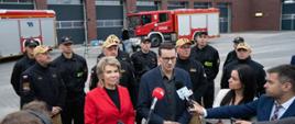 Zdjęcie przedstawia Pana Prezesa Rady Ministrów Pana Mateusza Morawieckiego podczas briefingu prasowego. Wokół zgromadzeni strażacy oraz senator RP Pani Dorota Tobiszowska. W tle samochody pożarnicze oraz nowa strażnica KM PSP w Rudzie Śląskiej.