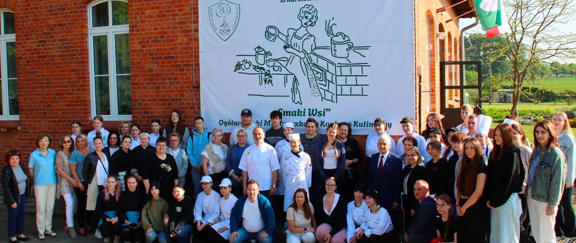 Grupowe zdjęcie uczestników i organizatorów wydarzenia plenerowego na tle budynku i dużego banera informacyjnego.