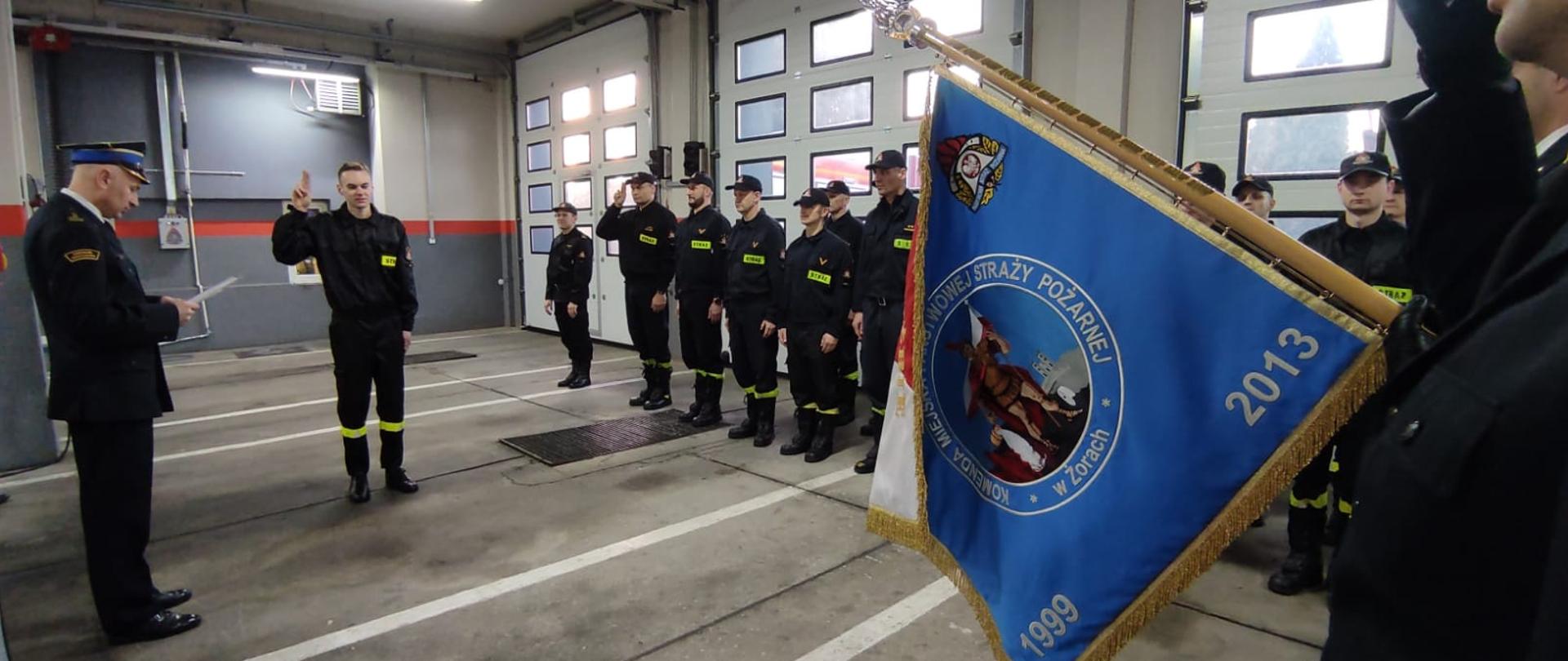 Strażacy stojący w dwuszeregu, na środku ślubujący strażak, w lewej części zdjęcia Komendant, w prawej jego części sztandar Komendy.