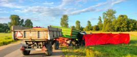 Przewrócony na lewy bok ciągnik rolniczy, znajdujący się poza pasem drogi gminnej z podczepionymi dwiema przyczepami rolniczymi stojącymi na drodze, obie do pełna załadowane zbożem. Do ciągnika rolniczego przylega rozstawiony przez straż pożarną czerwony parawan. Zdjęcie wykonano w słoneczny dzień z drogi gminnej.