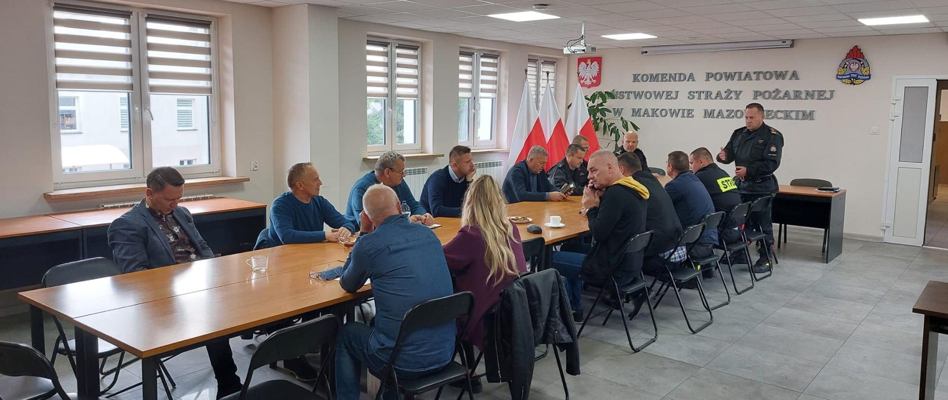 Strażacy oraz samorządowcy siedzą naprzeciwko siebie przy stole na świetlicy. Na ścianie znajduje się napis "Komenda Powiatowa Państwowej Straży Pożarnej w Makowie Mazowieckim". Jeden z nich stoi i przemawia.