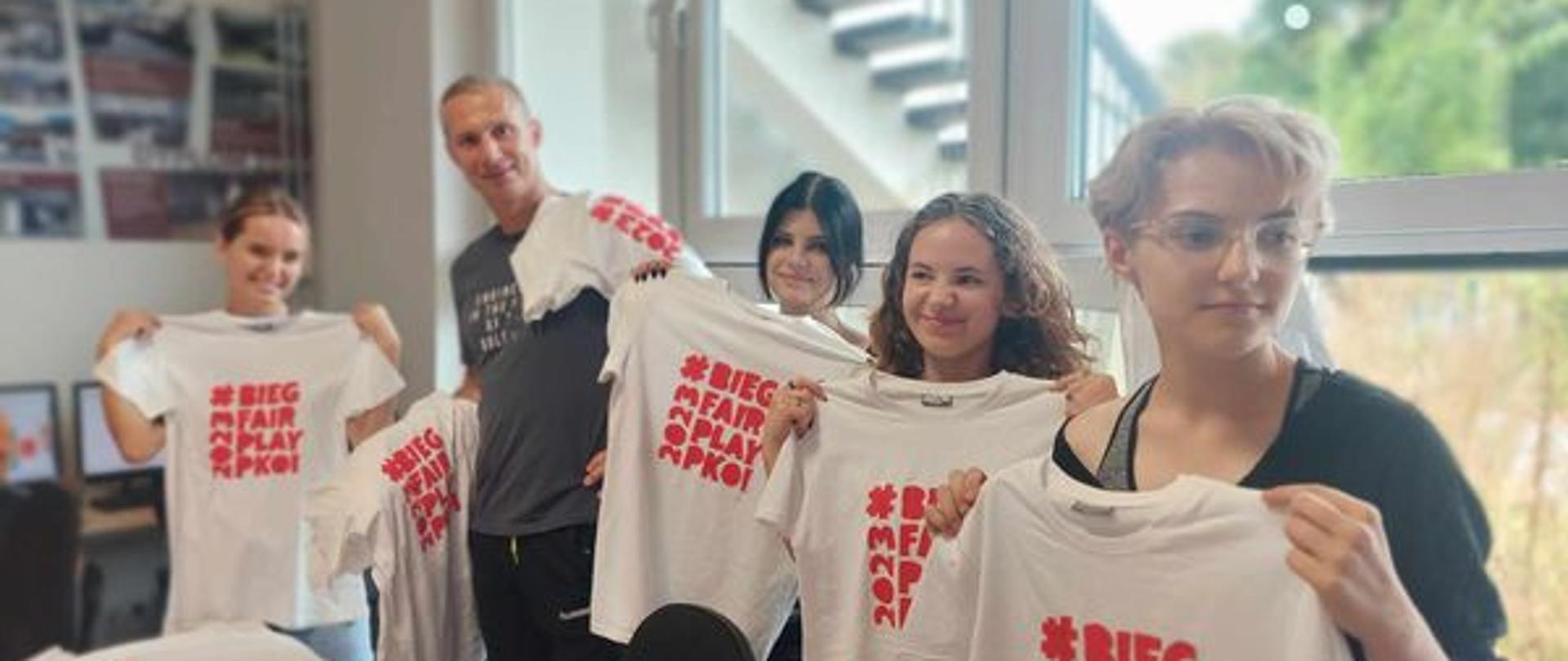 W pracowni tkaniny artystycznej PLSP w Lublinie uczennice i nauczyciel Tomasz Krasowski trzymają w rękach rozłożone białe koszulki z czerwonym napisem "Bieg Fair Play PKOL".