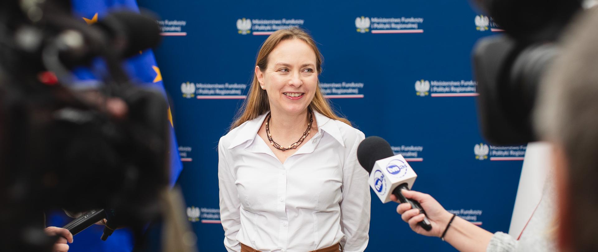 Minister Katarzyna Pełczyńska-Nałęcz stoi pod ścianą nazwą ministerstwa na niebieskim tle.