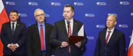 Czterech mężczyzn w garniturach stoi przed niebieską ścianą, za nimi po bokach polskie flagi, wśród nich minister Czarnek który mówi do stojącego na stojaku mikrofonu i trzyma w ręku czerwoną teczkę.