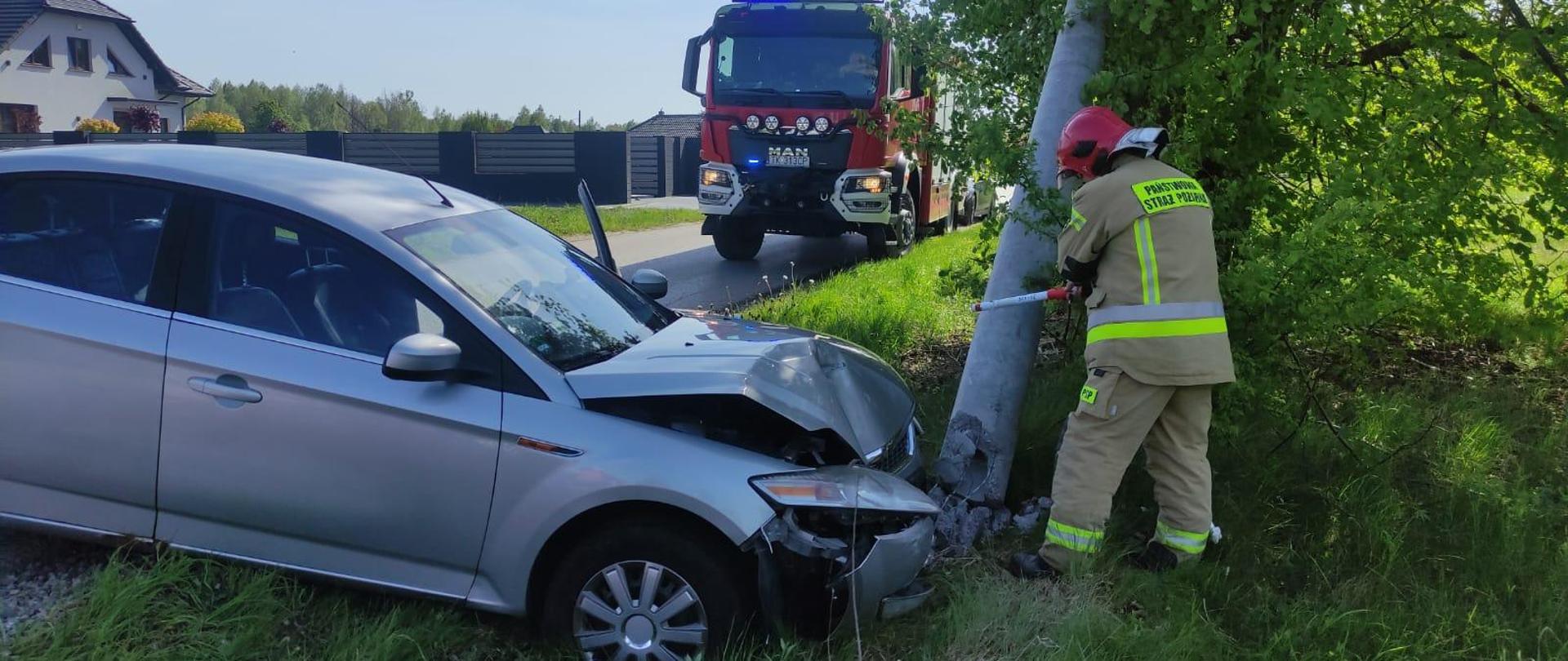 Zdjęcie przedstawia samochód osobowy stojący w rowie przodem oparty o betonowy słup. Zniszczony jest zarówno przód auta jak i słup oświetleniowy, w którym strażak detektorem sprawdza napięcie. W tle widać samochód pożarniczy.
