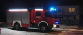 Obok posesji na drodze znajduje się pojazd pożarniczy z włączoną sygnalizacją świetlną.