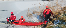 Troje strażaków wchodzi na staw pokryty lodem. Wszyscy ubrani w skafandry do pracy w wodzie i zabezpieczeni linkami oraz kapokami. Zabierają ze sobą sanie lodowe, za pomocą których podejmą osobę poszkodowaną z wody.