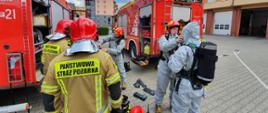 Zdjęcie przedstawia strażaków ćwiczących ratownictwo chemiczno - ekologiczne na placu Jednostki Ratowniczo - Gaśniczej.
W tle plac komendy, samochody oraz budynki mieszkalne.