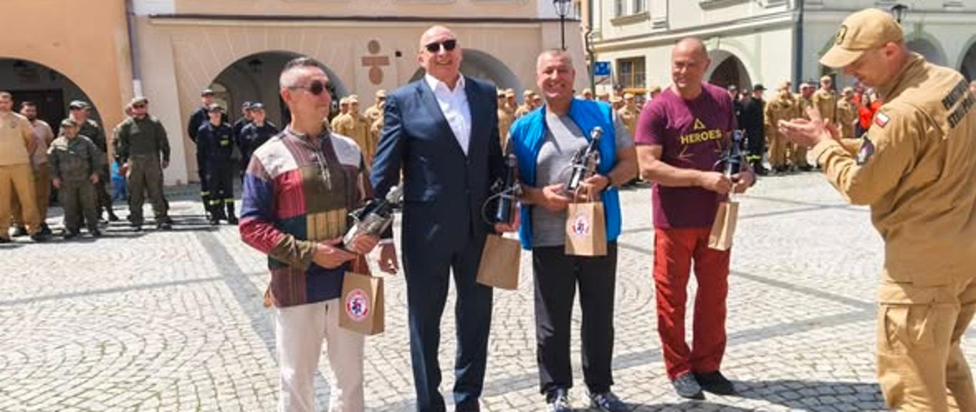 Oficjalne wręczenie podziękowań na brukowanym Placu Ratuszowym w Jeleniej Górze. W centrum kadru stoi czterech uśmiechniętych mężczyzn trzymających papierowe torebki z upominkami oraz butelki. Od lewej stoją: mężczyzna w jasnych spodniach i wzorzystej koszuli, mężczyzna w ciemnym garniturze i okularach przeciwsłonecznych, mężczyzna w sportowej niebieskiej kamizelce oraz mężczyzna w bordowej koszulce i czerwonych spodniach. Z prawej strony przodem do nich stoi strażak w piaskowym mundurze i czapce z daszkiem, który klaszcze w dłonie. W tle, przed zabytkowymi kamienicami z podcieniami, w równych rzędach stoją zgrupowane formacje strażackie w ciemnych i piaskowych mundurach.