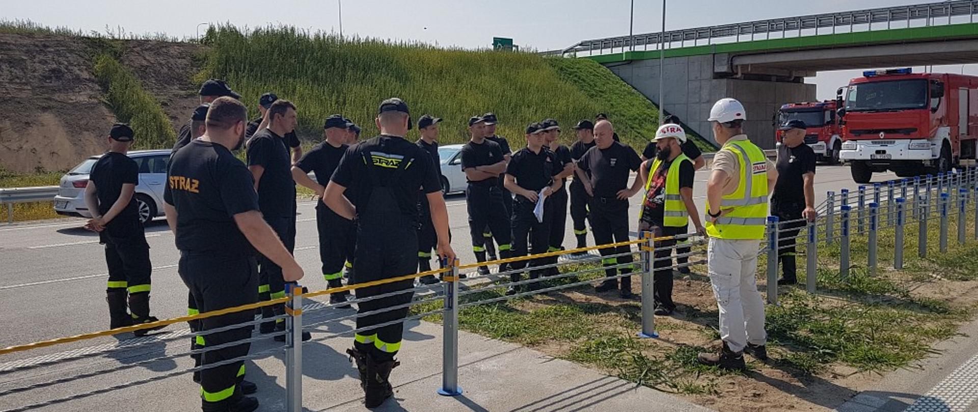 Na zdjęciu widać strażaków i pracowników firmy Strabag stojących przy barierkach oddzielających pasy drogi, w tle pojazdy pożarnicze