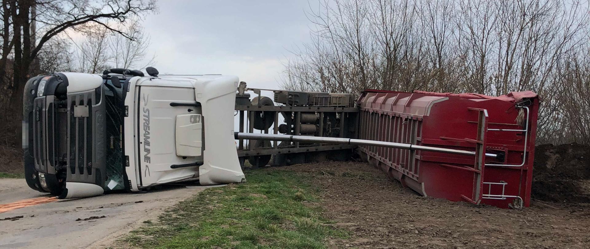 Zdjęcie zostało wykonane w ciągu dnia w godzinach popołudniowych. Zdjęcie przedstawia wywrócony samochód ciężarowy z naczepą samowyładowczą, który leży na lewym boku. Ciągnik siodłowy znajduje się na drodze natomiast jego naczepa leży na zaoranym polu . W tle widoczne drzewa oraz linie energetyczne.