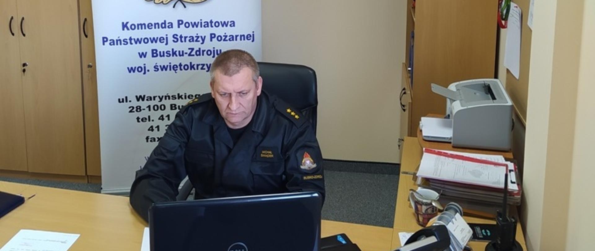 Na zdjęciu strażak w ubraniu dowódczo-sztabowym w kolorze czarnym siedzi przy biurku przed laptopem,za nim rollup przedstawiający logo straży pożarnej i adres komendy.