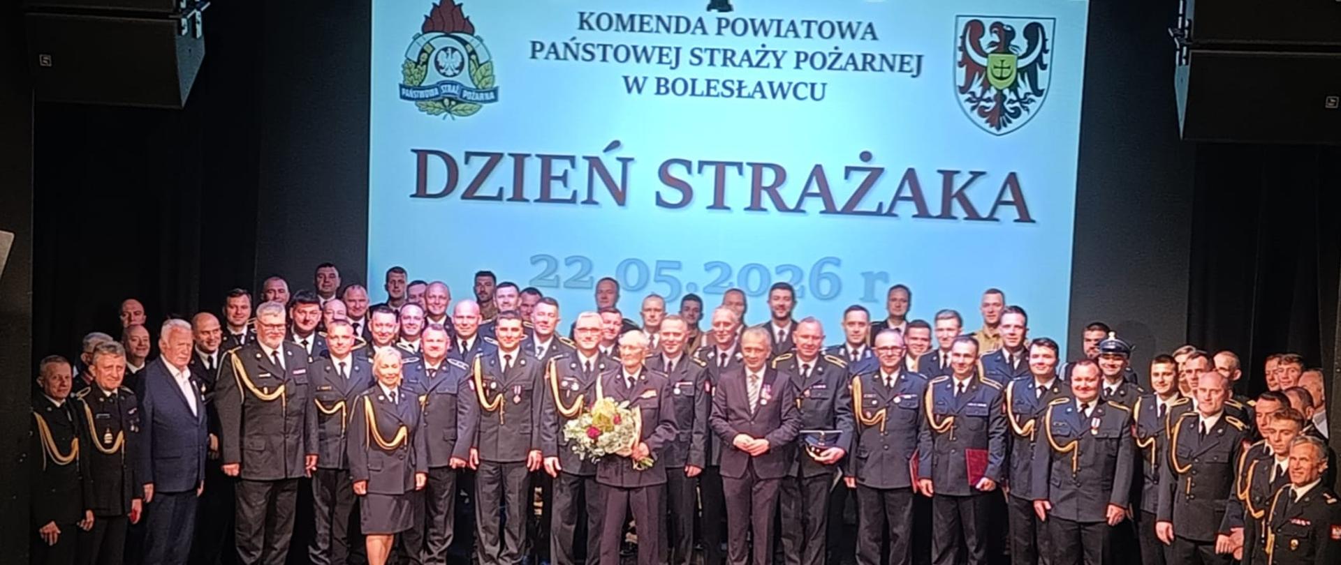Pamiątkowe zdjęcie grupowe kilkudziesięciu strażaków w galowych mundurach na scenie. W tle wyświetlany jest duży napis „KOMENDA POWIATOWA PAŃSTWOWEJ STRAŻY POŻARNEJ W BOLESŁAWCU. DZIEŃ STRAŻAKA. 22.05.2026 r.” oraz logo formacji i herb. Na środku stoi wyróżniony strażak trzymający bukiet kwiatów.