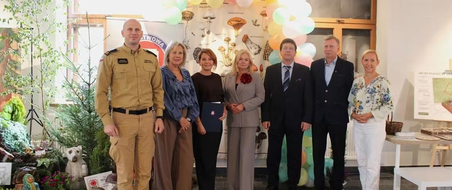 Grupa siedmiu osób pozuje do zdjęcia w sali udekorowanej kolorowymi balonami i elementami wystawy przyrodniczej.