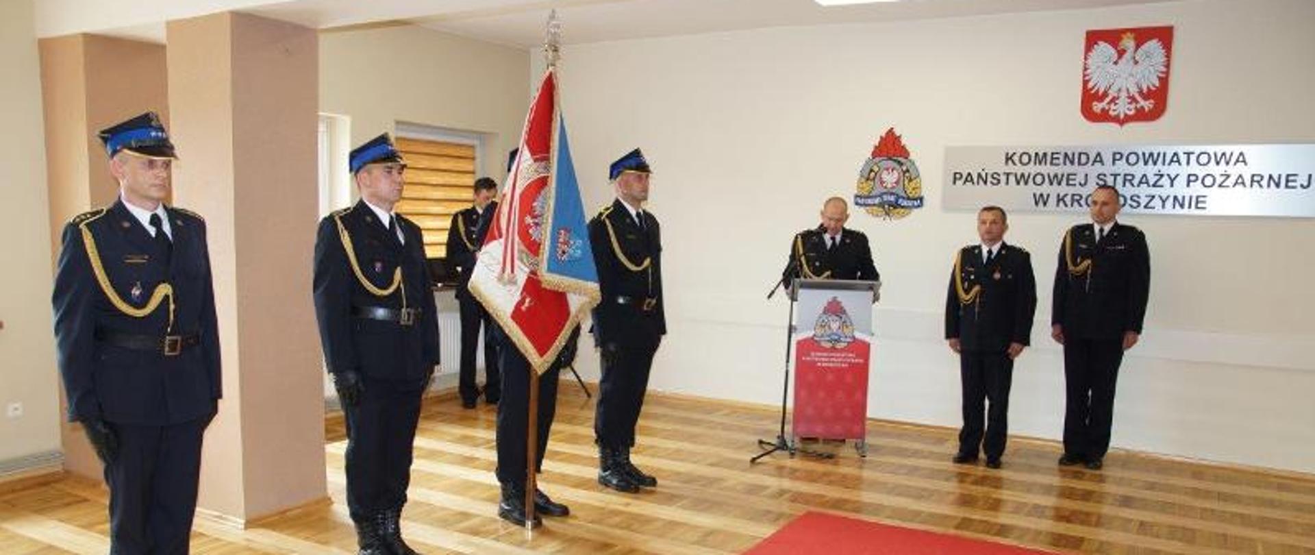 Podczas ceremonii pożegnania komendanta powiatowego, który przechodzi na zaopatrzenie emerytalne przemawia dowódca jednostki. Podczas wystąpienia przy mównicy przedstawia życiorys komendanta. Obok mównicy stoi komendant powiatowy, który zdaje swoje obowiązki oraz zastępca komendanta. Po lewej stronie funkcjonariusze pocztu sztandarowego trzymają sztandar komendy. Na podłodze w świetlicy komendy przed sztandarem rozłożono czerwony dywan 