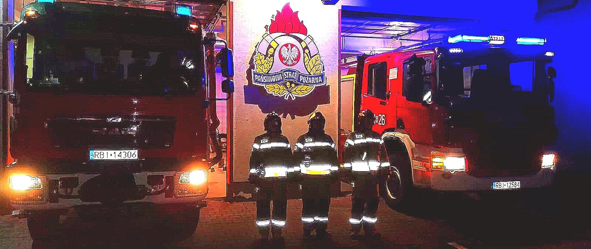 Na zdjęciu widać trzech strażaków PSP w ubraniach specjalnych którzy stoją przed garażem JRG w Ustrzykach Dolnych, za plecami na ścianie wisi logo PSP a z obu ich stron wystawione są samochody pożarnicze które mają włączone światła mijania oraz światła alarmowe. 