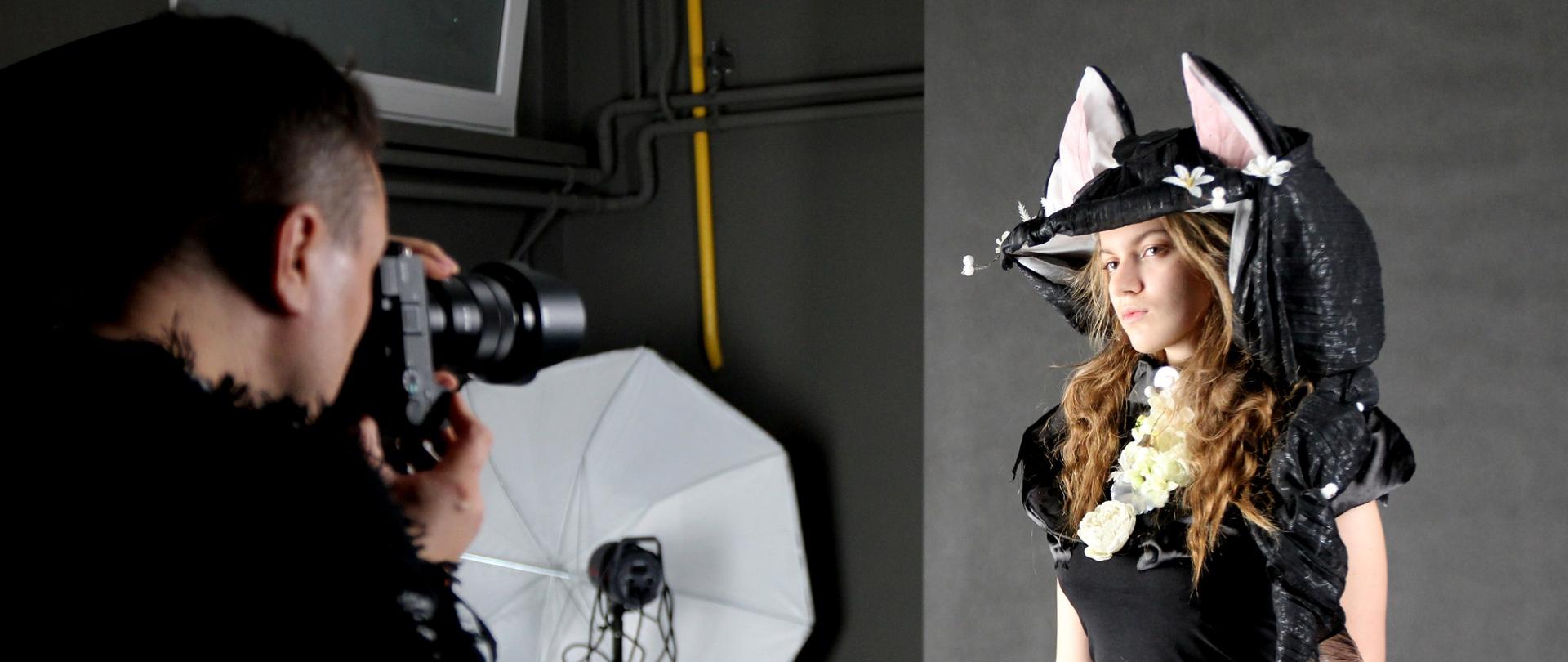 Fotograf wykonuje portret młodej dziewczyny w studiu fotograficznym. Modelka stoi na tle szarego tła, ubrana w czarną bluzkę i dżinsy, ma na głowie artystyczne nakrycie z czarną tkaniną, kwiatami i ozdobnymi uszami. Obok widoczna lampa studyjna z softboxem.