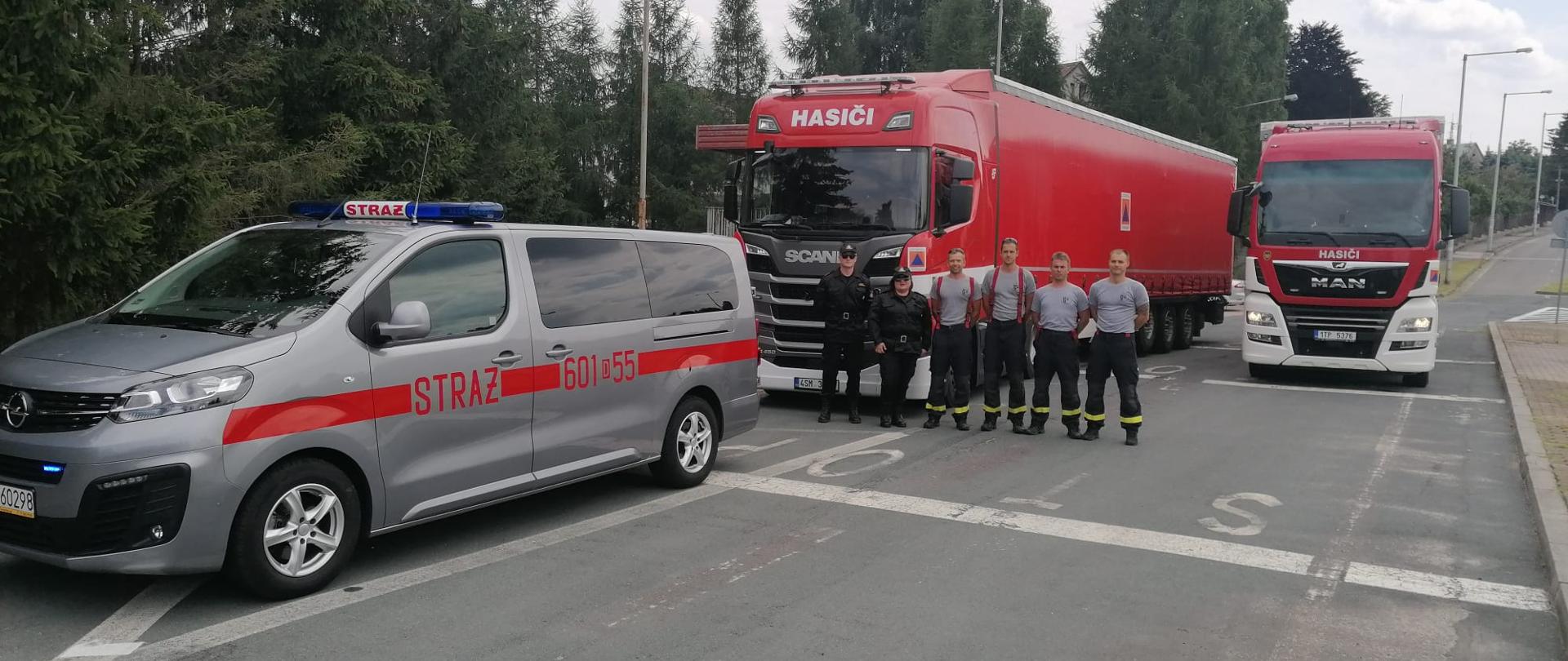 Samochód typu bus z numerami operacyjnymi 601D55. Za nim w szeregu dwójka funkcjonariuszy PSP w mundurach służbowych (mężczyzna i kobieta). Obok nich czerech strażaków z Czech. Za nimi dwa samochody ciężarowe TIR.