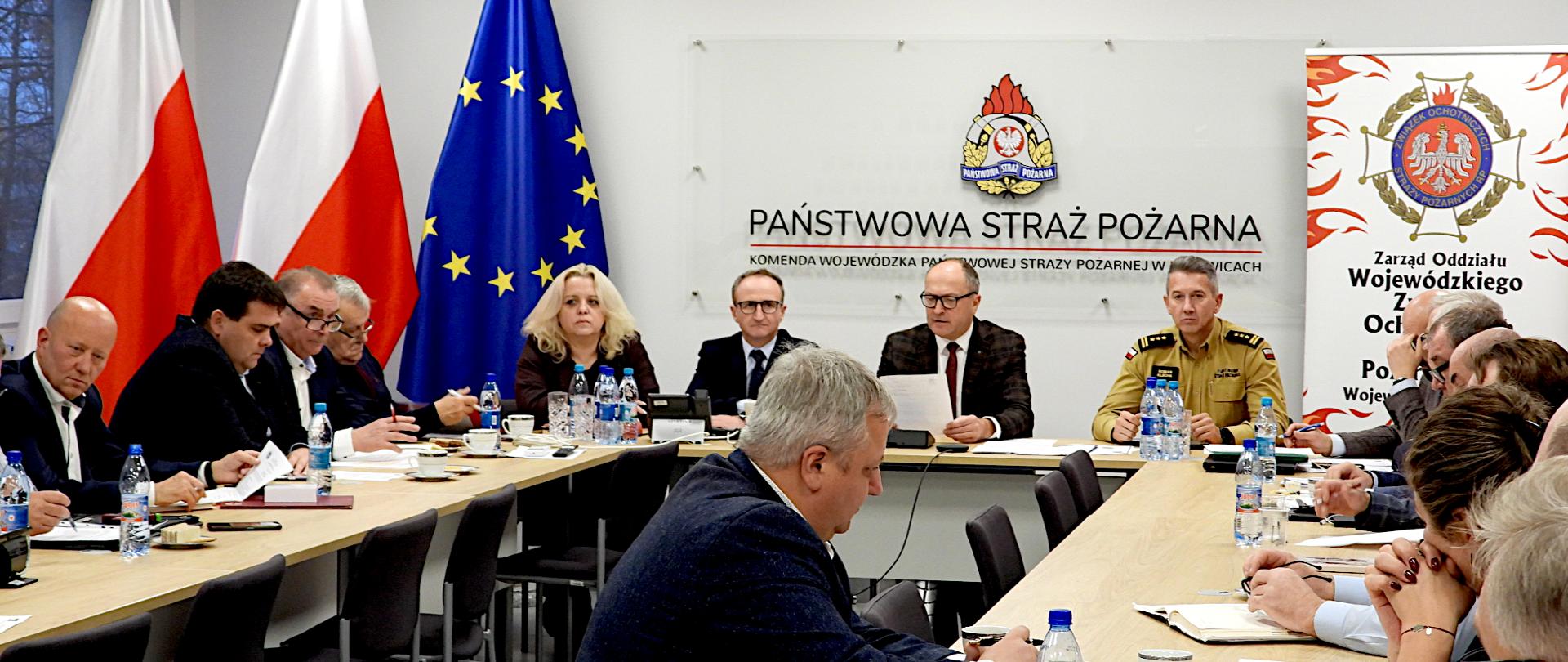 Na zdjęciu widać spotkanie posiedzenie Prezydium Zarządu Oddziału Wojewódzkiego Związku Ochotniczych Straży Pożarnych RP województwa śląskiego w sali Komendy Wojewódzkiej Państwowej Straży Pożarnej w Katowicach. Przy długim stole w kształcie prostokąta siedzi kilkanaście osób – głównie mężczyźni w garniturach, a także jedna kobieta. Na środku stołu, po stronie głównej, siedzi mężczyzna w okularach i garniturze, który trzyma kartkę i prawdopodobnie prowadzi zebranie. Obok niego znajduje się mężczyzna w piaskowym mundurze służbowym - Jest to st. bryg. Roman Klecha - Zastępca Śląskiego Komendanta Wojewódzkiego. Na ścianie za nimi widnieje duże logo Państwowej Straży Pożarnej oraz napis „Komenda Wojewódzka Państwowej Straży Pożarnej w Katowicach”. Obok stoi roll-up z emblematem Ochotniczych Straży Pożarnych RP i napisem o wojewódzkim zarządzie związku. Z lewej strony sali widać flagi Polski i Unii Europejskiej. Na stołach leżą notatniki, dokumenty, długopisy, filiżanki z kawą i butelki z wodą. 