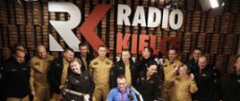 Na zdjęciu widzimy grupę strażaków w umundurowaniu służbowym piaskowym w studiu Radia Kielce. Oprócz mikrofonów i sprzętu radiowego, w tle na ścianie za grupą ludzi widzimy napis Radio Kielce