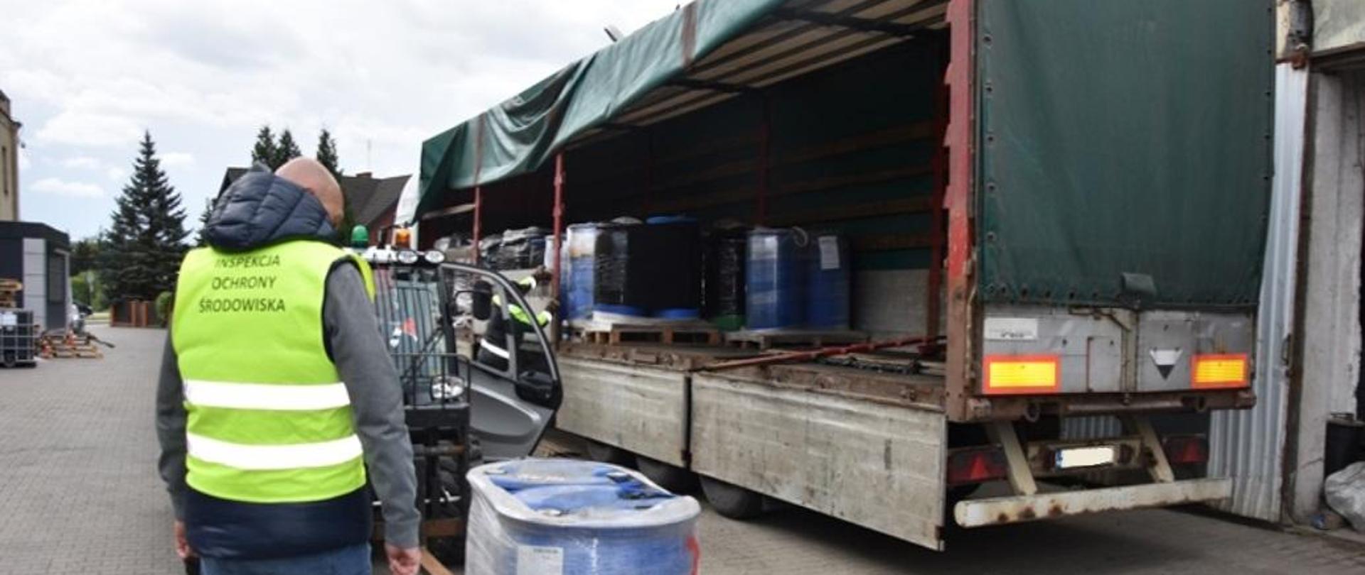 Pracownik Wojewódzkiego Inspektoratu Ochrony Środowiska w Warszawie kontroluje usuwanie niebezpiecznych odpadów. Po jego prawej stronie stoją na palecie dwie beczki o pojemności 200 l każda. W tle znajduje się samochód ciężarowy oraz wózek widłowy.