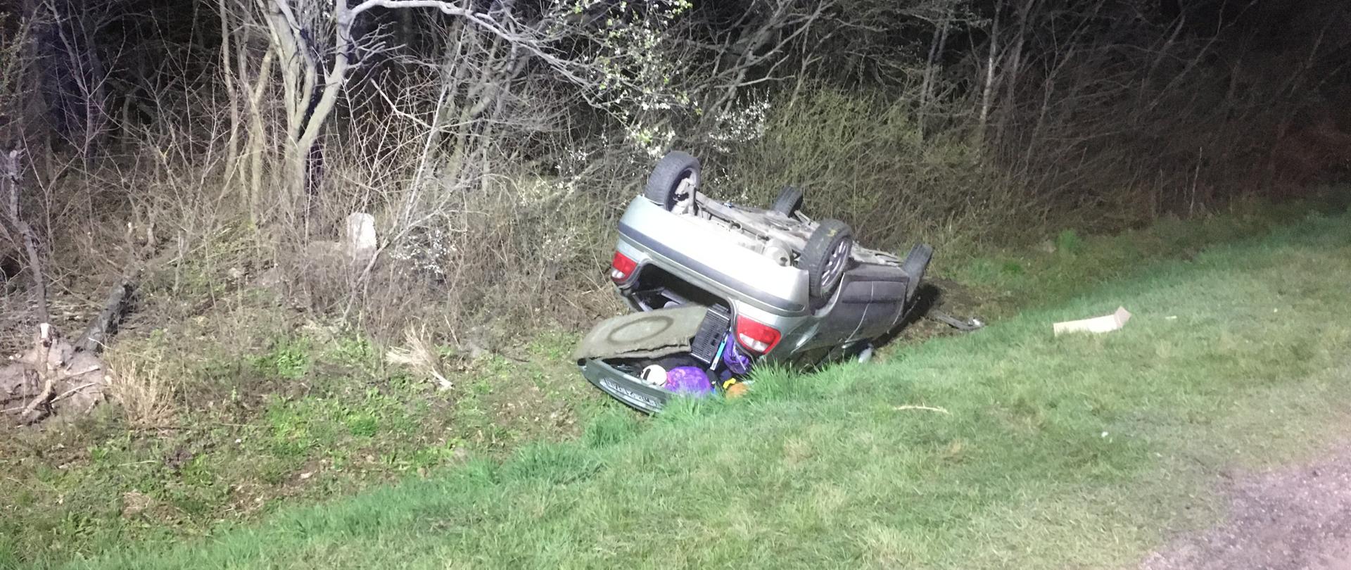 Srebrny samochód lezy na dachu poza drogą z otwarta klapa bagażnika widoczne elementy wyposażenia samochodu