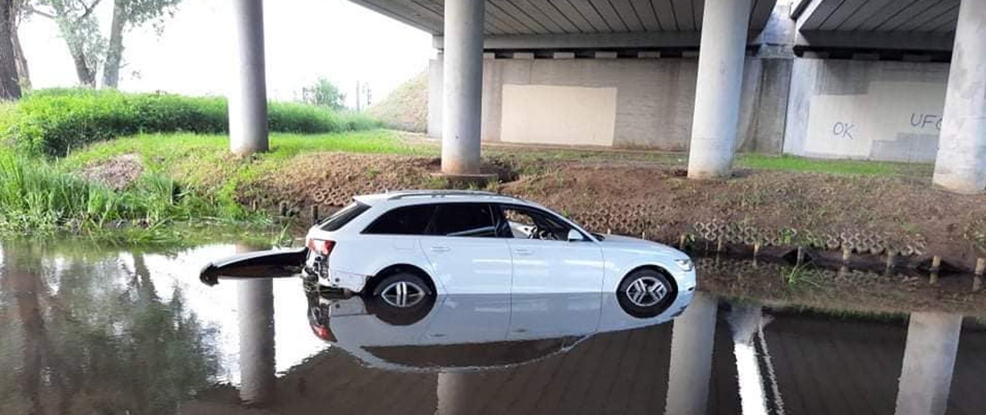 Na środku zdjęcia widoczny jest biały samochód osobowy Audi A6 z uszkodzonym tylnym zderzakiem. Pojazd znajduje się w rzece. Za pojazdem widoczny drugi brzeg porośnięty trawą oraz widoczne betonowe umocnienia brzegów rzeki. W dalszym planie widoczne szare betonowe słupy wspierające wiadukt znajdujący się nad samochodem. W lewym górnym rogu widoczne w oddali drzewa.