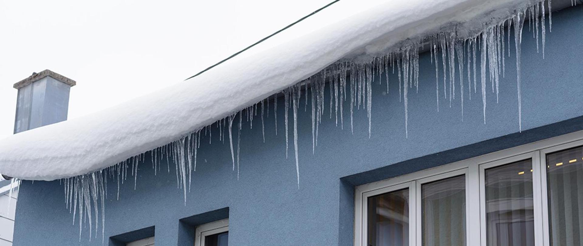 Zdjęcie przedstawia zalegający na dachu budynku śnieg i zwisające sople lodowe