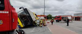 Na zdjęciu widać parking z kostki brukowej. Na środku znajduje się autobus komunikacji miejskiej. Jest pochylony. Niebo zachmurzone. Widać samochody straży pożarnej w kolorze czerwonym. Na parkingu latarnie oświetleniowe. Przy samochodach ratownicy.