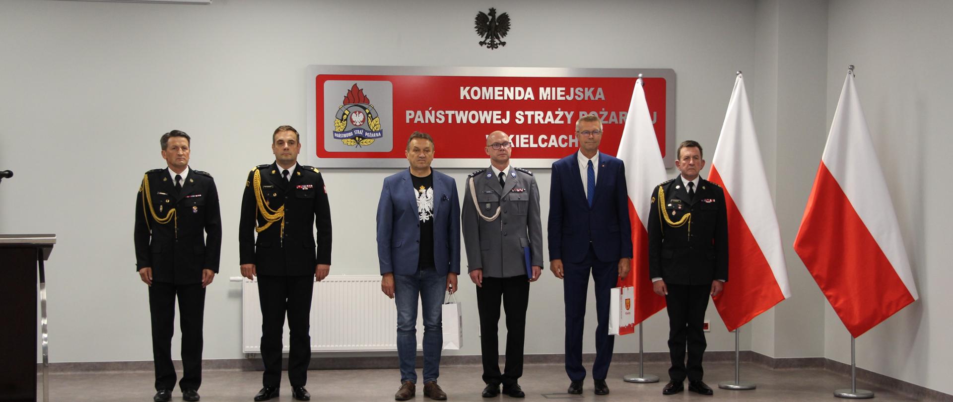 Zdjęcie przedstawia zaproszonych gości. Osoby te stoją w rzędzie na tle napisu Komenda Miejska PSP w Kielcach