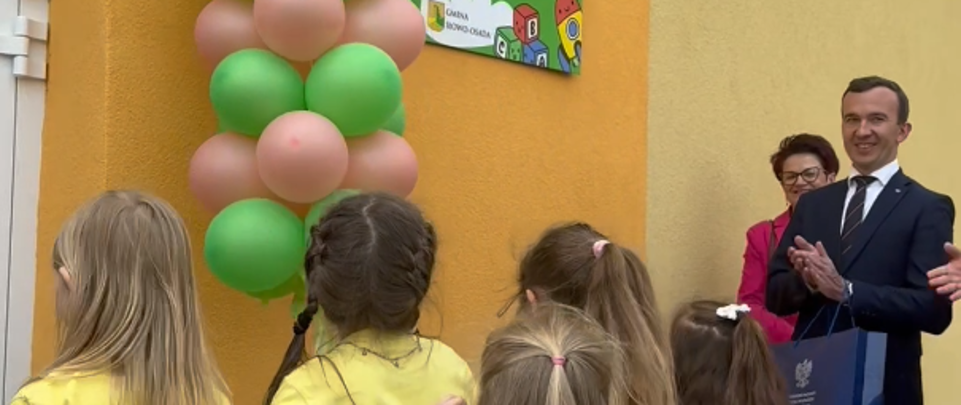 mężczyzna w garniturze klaszcze na widok odsłoniętego baneru na tle ściany budynku. Widać balony oraz dzieci