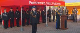 Na zdjęciu widzimy Komendanta Wojewódzkiego PSP podczas przemówienia przy mównicy oraz w tle pod namiotem zaproszonych gości i strażaków.ch.