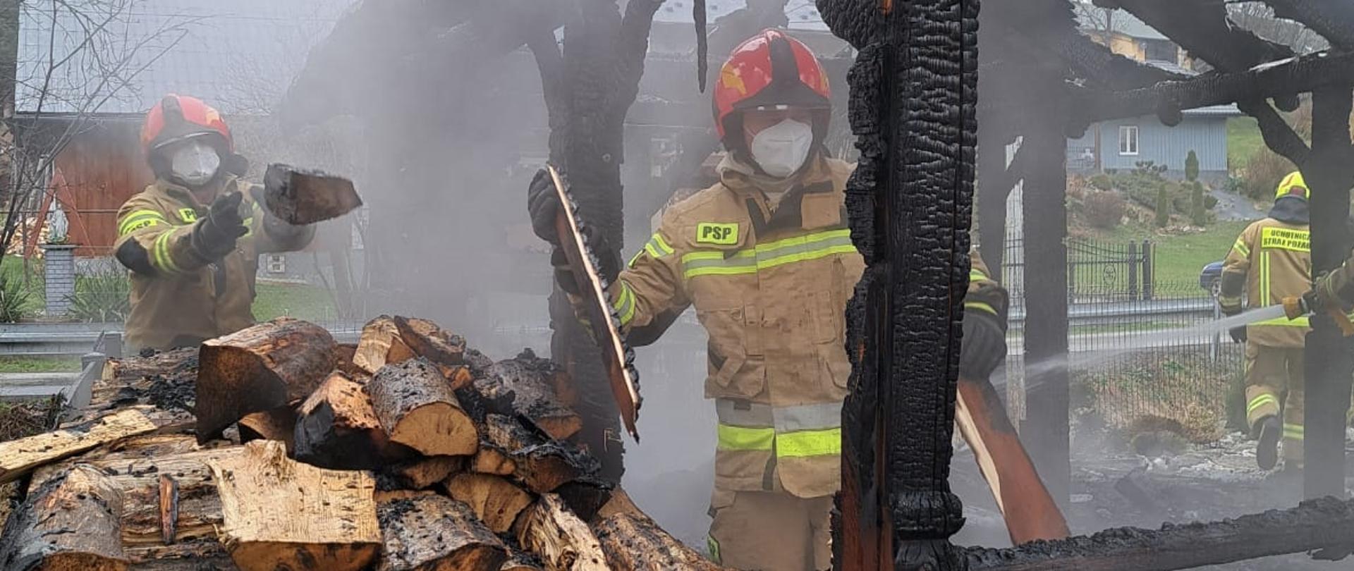 Kolorowa fotografia wykonana w pochmurny dzień. Przedstawia strażaków ubranych w ubrania specjalne w hełmach i maseczkach pracujących przy rozbiórce spalonej altany. W tle budynki mieszkalne.