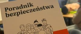 Okładka przedstawia ilustrację grupy osób, w tym rodzinę z dziećmi, psa oraz przedstawicieli służb mundurowych, na tle biało-czerwonej kolorystyki nawiązującej do barw narodowych. W tle zdjęcia znajduje się rozmyty baner z napisem „Wojewódzka Stacja Sanitarno-Epidemiologiczna w Białymstoku”. W pomieszczeniu widać również siedzące osoby, co sugeruje spotkanie informacyjne lub szkolenie. Całość wskazuje na wydarzenie związane z edukacją w zakresie bezpieczeństwa i zdrowia publicznego.