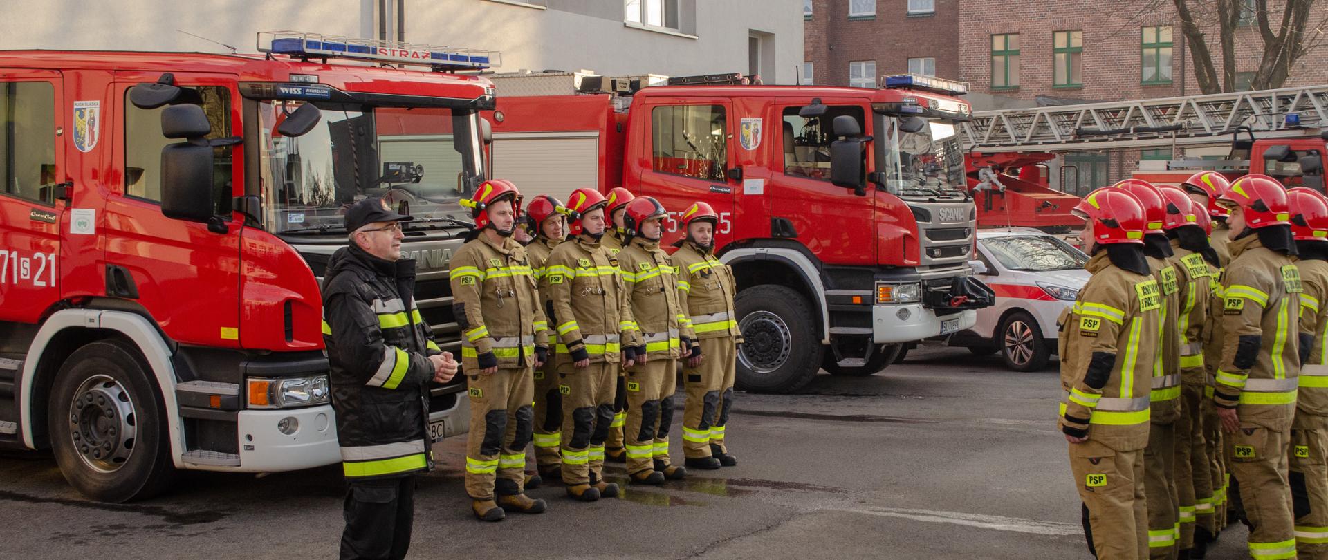 Na pierwszym planie widoczny jest idący bryg. Sławomir Gajda umundurowany w ubranie dowódczo- sztabowe. Na drugim planie znajdują się strażacy umundurowani w ubranie bojowe tworząc dwa rzędy zwrócone do siebie twarzami. Na drugim planie znajdują się samochody pożarnicze. W tle usytuowany jest budynek KM PSP w Rudzie Śląskiej.