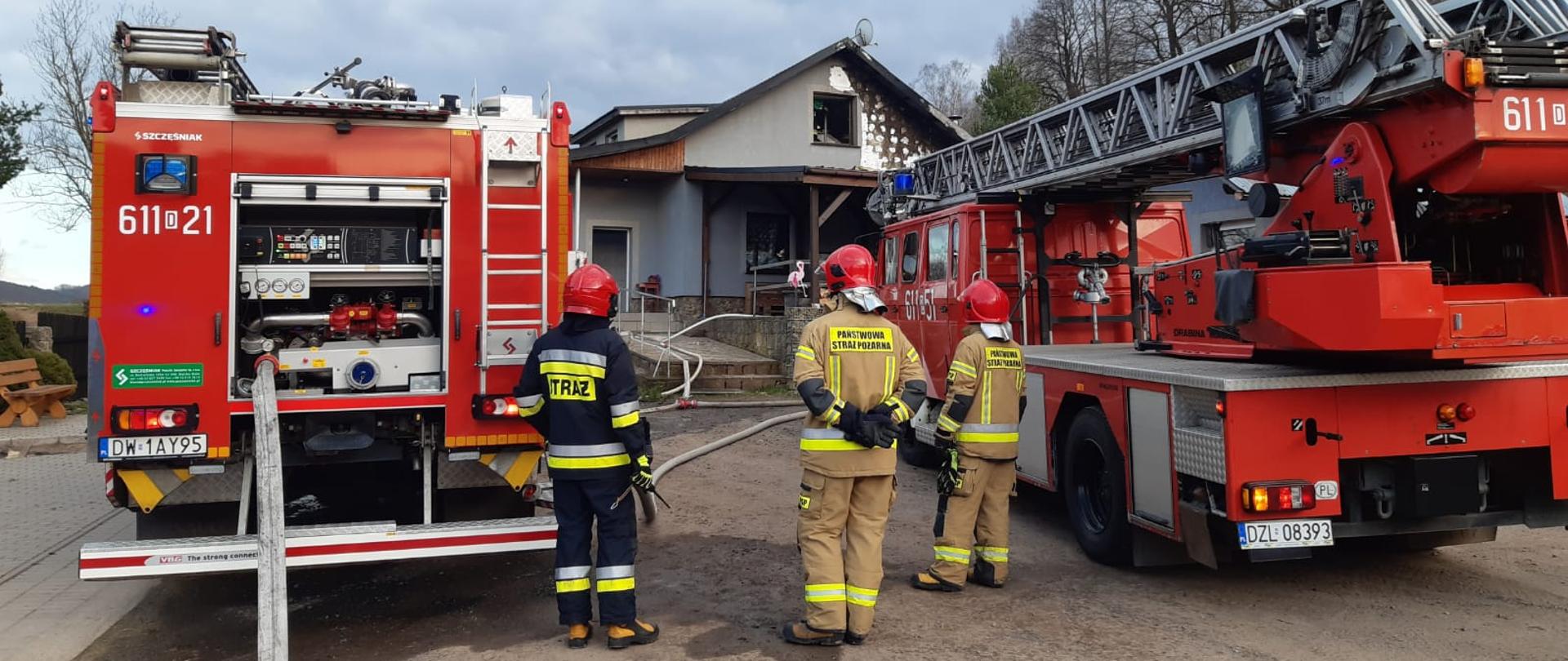 wozy strażackie, pośród nich trzech umundurowanych strażaków w czerwonych hełmach, w tle budynek jednorodzinny