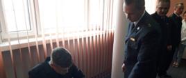 Na zdjęciu widać siedzącego strażaka, który siedzi przed stołem i podpisuje dokument. Po prawej funkcjonariusz w mundurze wyjściowym. Za strażakami zasłona i okno.