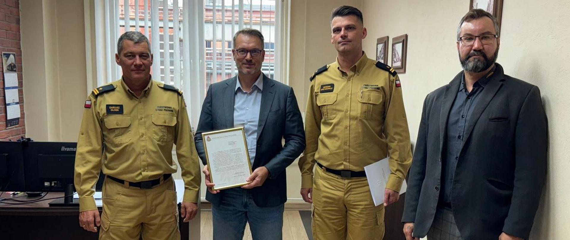 Na zdjęciu znajduje się Komendant Miejski PSP w Elblągu, Zastępca Dowódcy JRG w Pruszczu Gdańskim oraz Wiceprezes firmy Stokota i kierownik jednego z działów firmy. Funkcjonariusze PSP wręczają Panu Wiceprezesowi podziękowania od Komendanta Głównego PSP za prezentację cystern firmy Stokota. Mężczyźni znajdują się w gabinecie Wiceprezesa firmy Stokota.