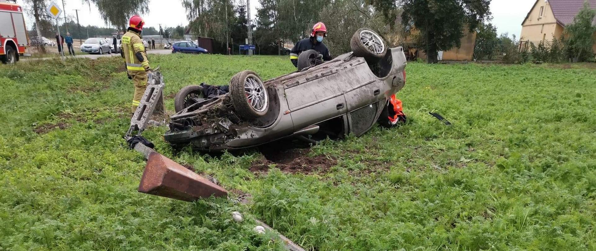 Samochód w polu leży przewrócony na dachu. W tyle dwóch strażaków działających przy akcji. Przed samochodom stoi złamany słup energetyczny.
