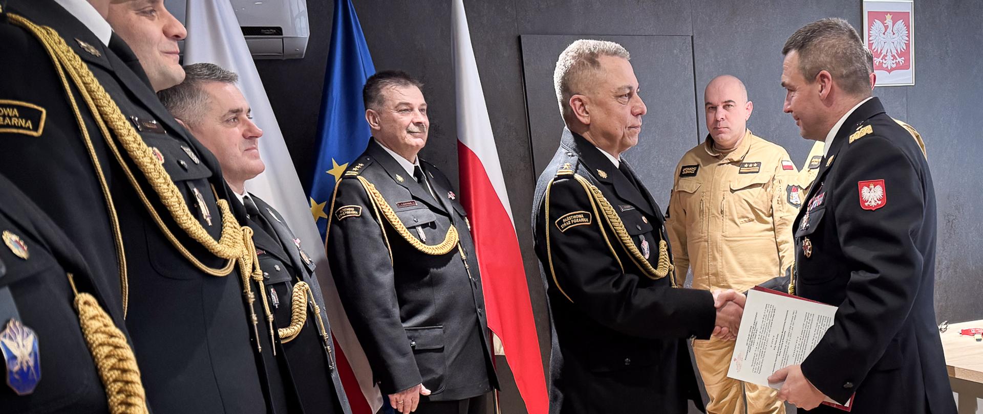 Moment wręczenia dokumentu. Podkarpacki komendant wojewódzki PSP nadbryg. Tomasz Baran podaje rękę bryg. Piotrowi Guzkowi, przekazując mu akt mianowania na stanowisko naczelnika. Obaj oficerowie są w pełnym umundurowaniu wyjściowym. W tle pozostali uczestnicy uroczystości.