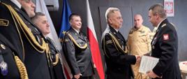 Moment wręczenia dokumentu. Podkarpacki komendant wojewódzki PSP nadbryg. Tomasz Baran podaje rękę bryg. Piotrowi Guzkowi, przekazując mu akt mianowania na stanowisko naczelnika. Obaj oficerowie są w pełnym umundurowaniu wyjściowym. W tle pozostali uczestnicy uroczystości.