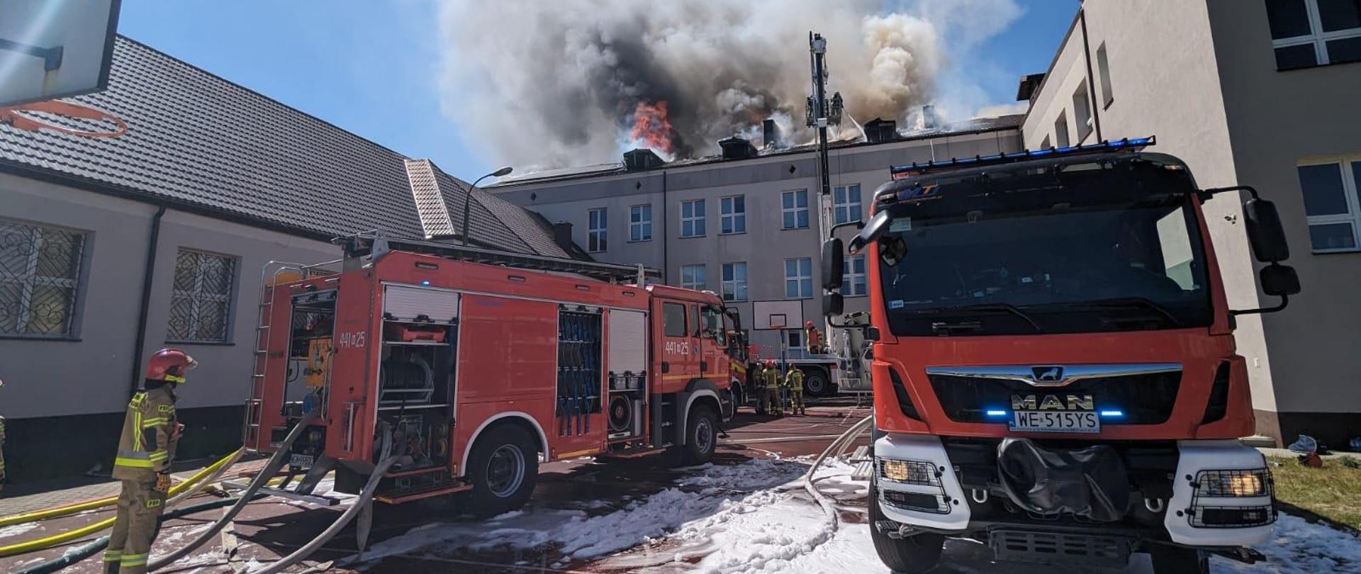 Na boisku szkolnym pokrytym białą pianą gaśniczą stoją dwa samochody gaśnicze i podnośnik hydrauliczny. Na boisku stoją strażacy. W koszu podnośnika jest dwóch strażaków, którzy leją wodę na widoczne zarzewia ognia na dachu budynku szkoły. Ponad dachem widać czarno-siwo-biały dym zmieszany z płomieniami wydobywającymi się z pod blachy. Niebo jest bezchmurne niebieskie. 