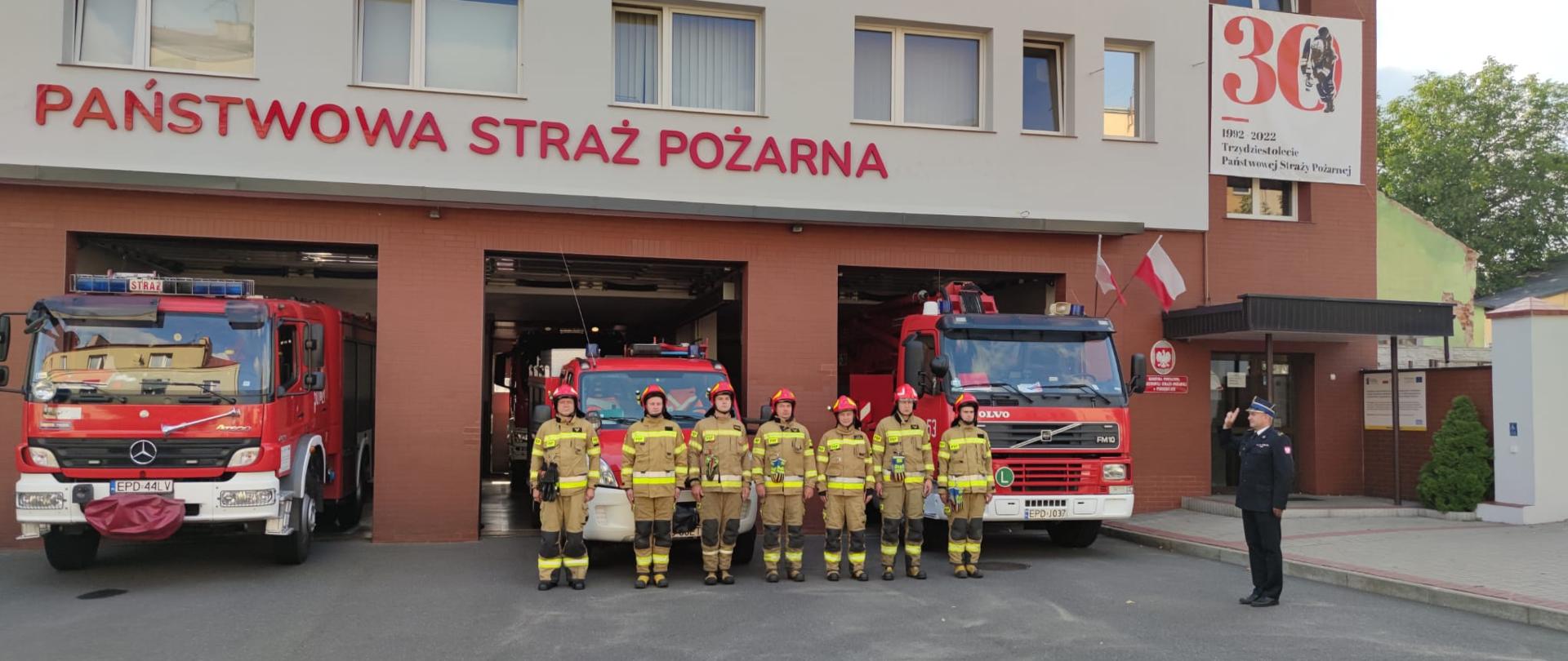 Przed budynkiem komendy częściowo wyprowadzone samochody. Przed nimi stoi 7 strażaków ubrani w hełmy i stroje koloru piaskowgo. Po prawo bokiem do strażaków na baczność stoi komendant powiatowy ubrany w czarny mundur strażacki , salutuje