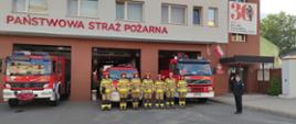 Przed budynkiem komendy częściowo wyprowadzone samochody. Przed nimi stoi 7 strażaków ubrani w hełmy i stroje koloru piaskowgo. Po prawo bokiem do strażaków na baczność stoi komendant powiatowy ubrany w czarny mundur strażacki , salutuje