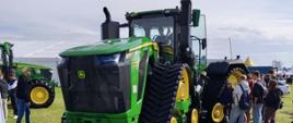 Duży zielony ciągnik John Deere na czterech gąsienicach stoi na trawie. Obok grupa młodzieży ogląda maszynę, w tle inne ciągniki i namioty wystawców.