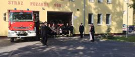 Z lewej strażacy w czarnych ubraniach koszarowych stojący w szeregu, z prawej Komendant Miejski PSP w Bydgoszczy wraz z Panią Poseł i Panią Radną, w środku kolejnych kilku strażaków w szeregu. Z lewej w tle przekazywany pojazd stojący częściowo w garażu JRG4.