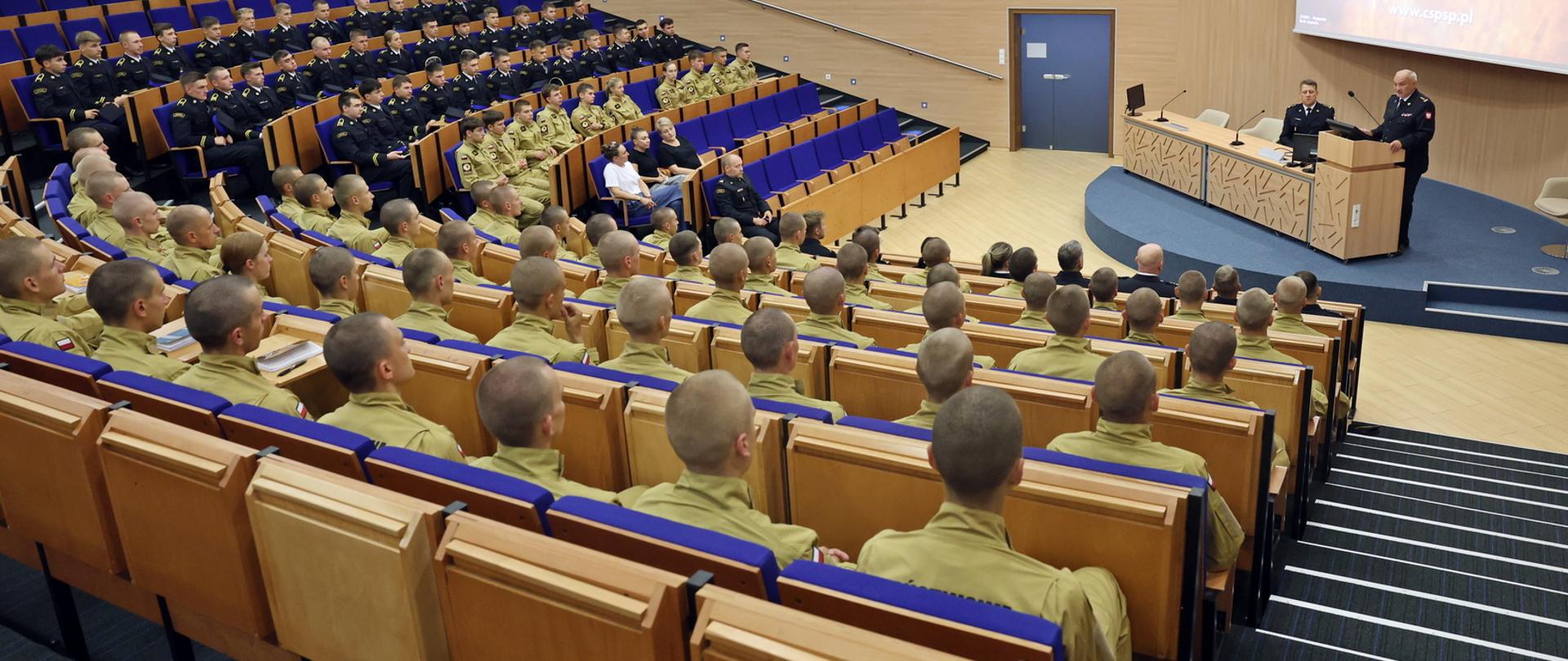 Widok z tyłu na siedzących w auli wykładowej CS PSP kadetów oraz funkcjonariuszy i pracowników cywilnych. W głębi na podeście stojąc przy mównicy stołu prezydialnego przemawia Komendant Centralnej Szkoły PSP