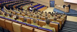 Widok z tyłu na siedzących w auli wykładowej CS PSP kadetów oraz funkcjonariuszy i pracowników cywilnych. W głębi na podeście stojąc przy mównicy stołu prezydialnego przemawia Komendant Centralnej Szkoły PSP