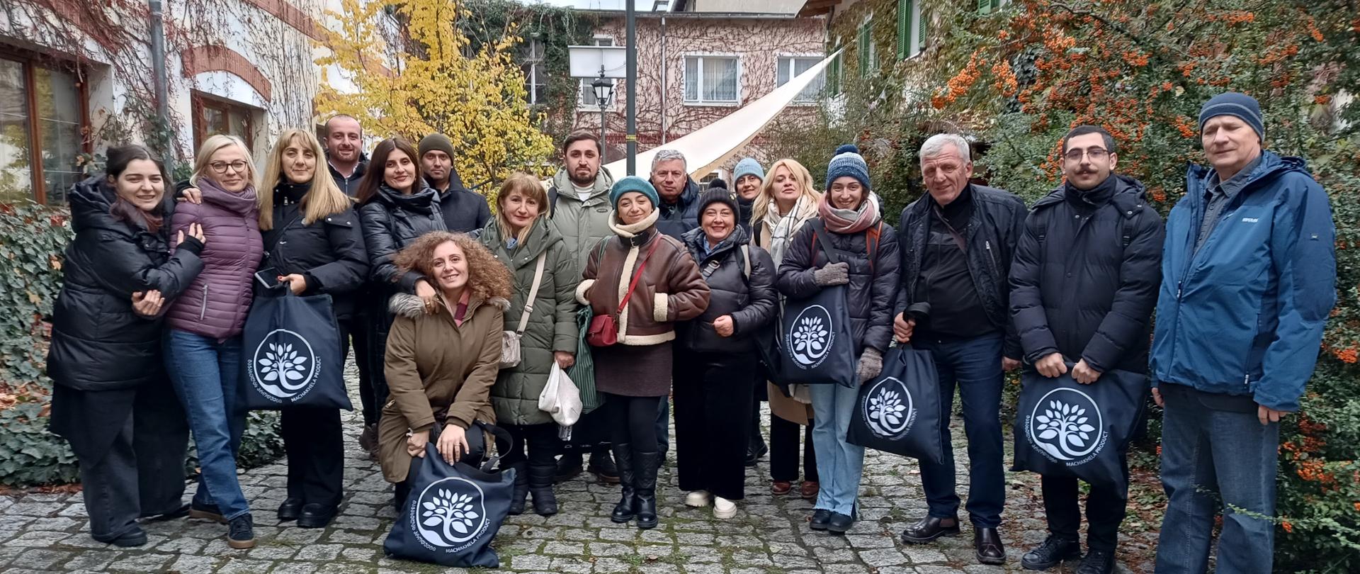 Grupa kobiet i mężczyzn pozuje do zdjęcia z torbami z napisem Machakhela Product, w tle obrośnięte poroślami budynki 