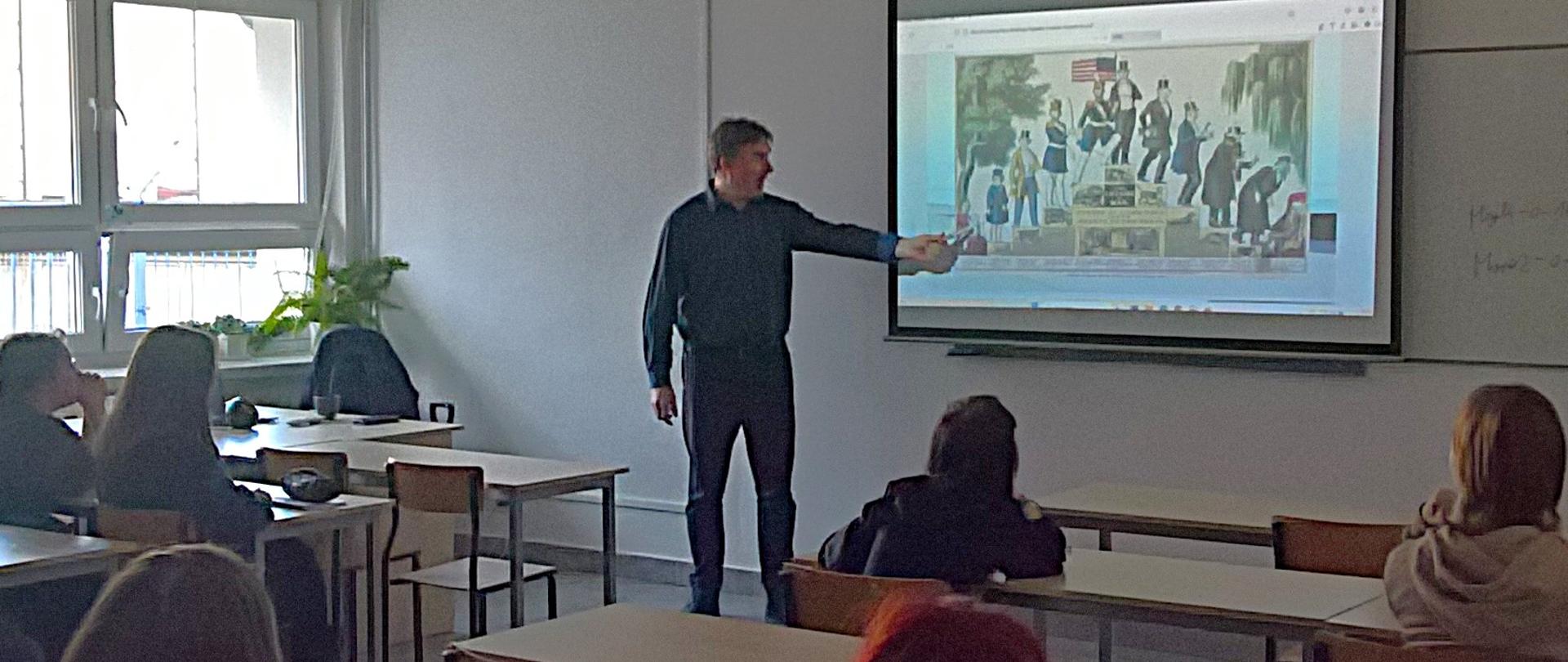 Sala lekcyjna, za oknami jasno, okna zasłonięte roletami. Mężczyzna stoi przy ekranie, wskazuje na wyświetlany na nim obraz. Osoby w ławkach, widziane z tyłu, patrzą w stronę ekranu